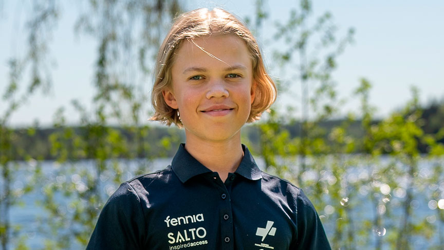 Aukusti Paakkala ulkona auringossa hymyilemässä kameralle. Taustalla vihreää kasvustoa ja vesistö.